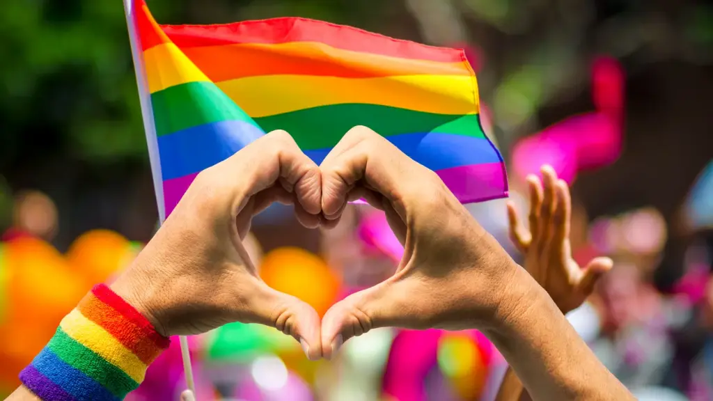 Mão fazendo coração com bandeira LGBTQIA+ ao fundo, representando inclusão e o debate sobre pink-washing.